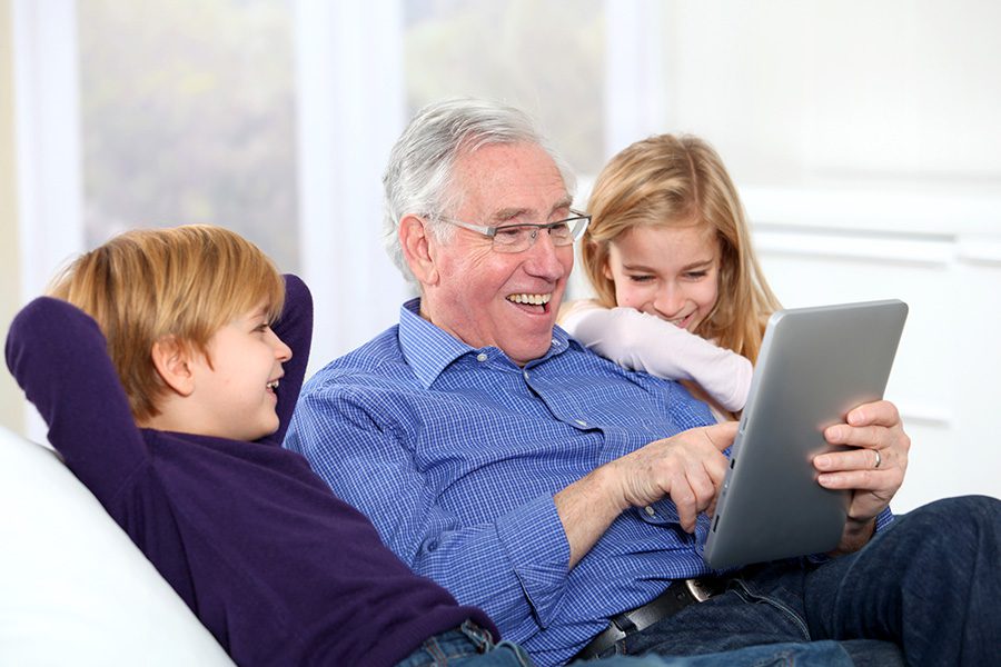 Client Center - A Grandfather is Sitting With His Grandchildren on a Sofa While Holding a Tablet and Smiling With Them