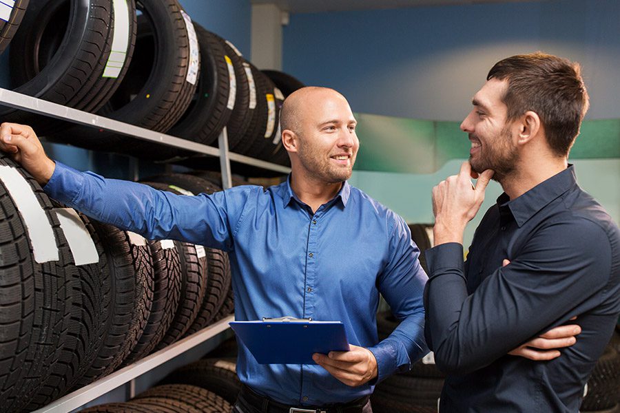 Business Insurance - Man Leaning on a Rack of Car Tires and Holding a Clipboard is Talking to a Male Customer About a Potential Sale at an Auto Store