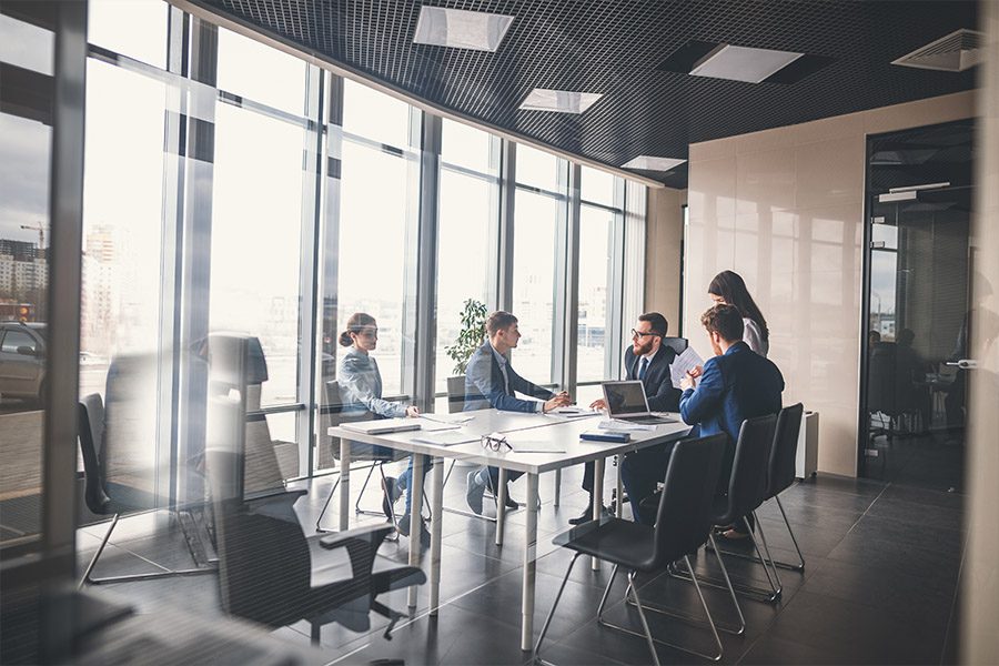 About - Business Professionals Sitting Around a Conference Table in a Modern Office with Large Windows and Light Coming in Behind Them